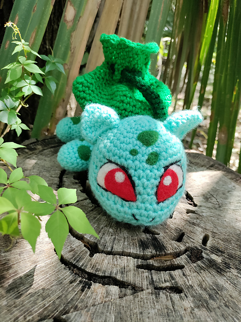 a crocheted bulbasaur sitting on a stump surrounded by greenery