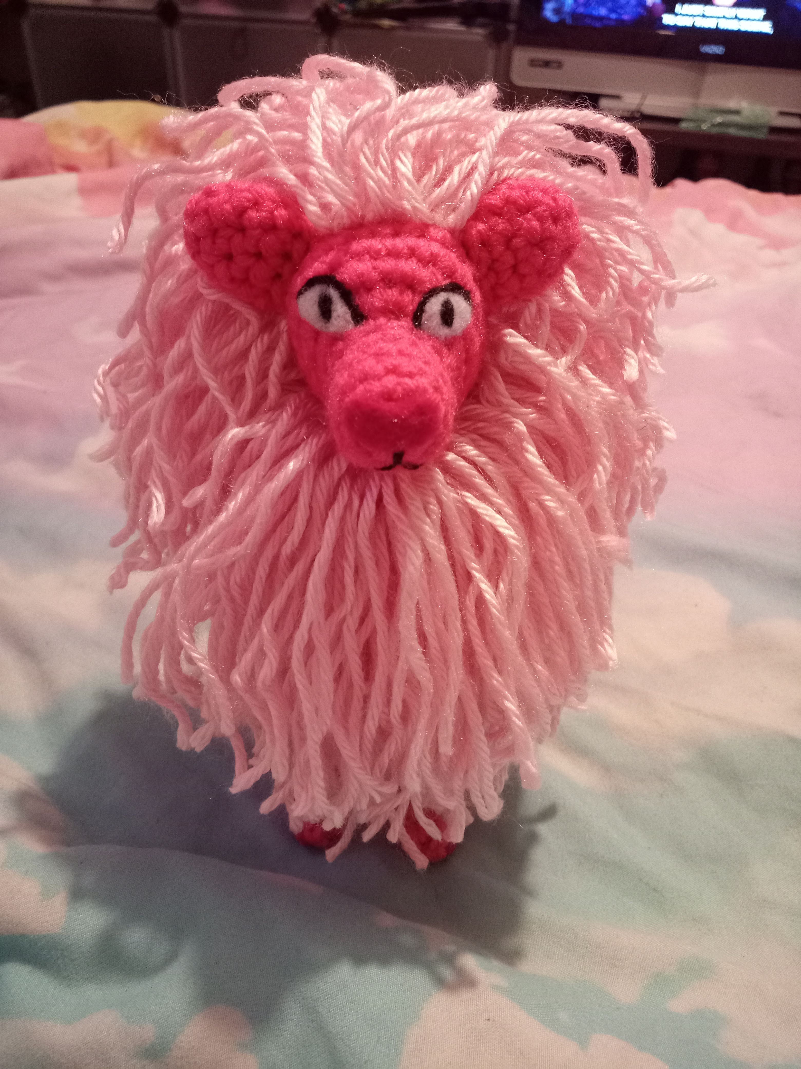 a front view of a crocheted Lion from Steven Universe standing on a cloudy sky comforter with unaltered yarn strings as the mane