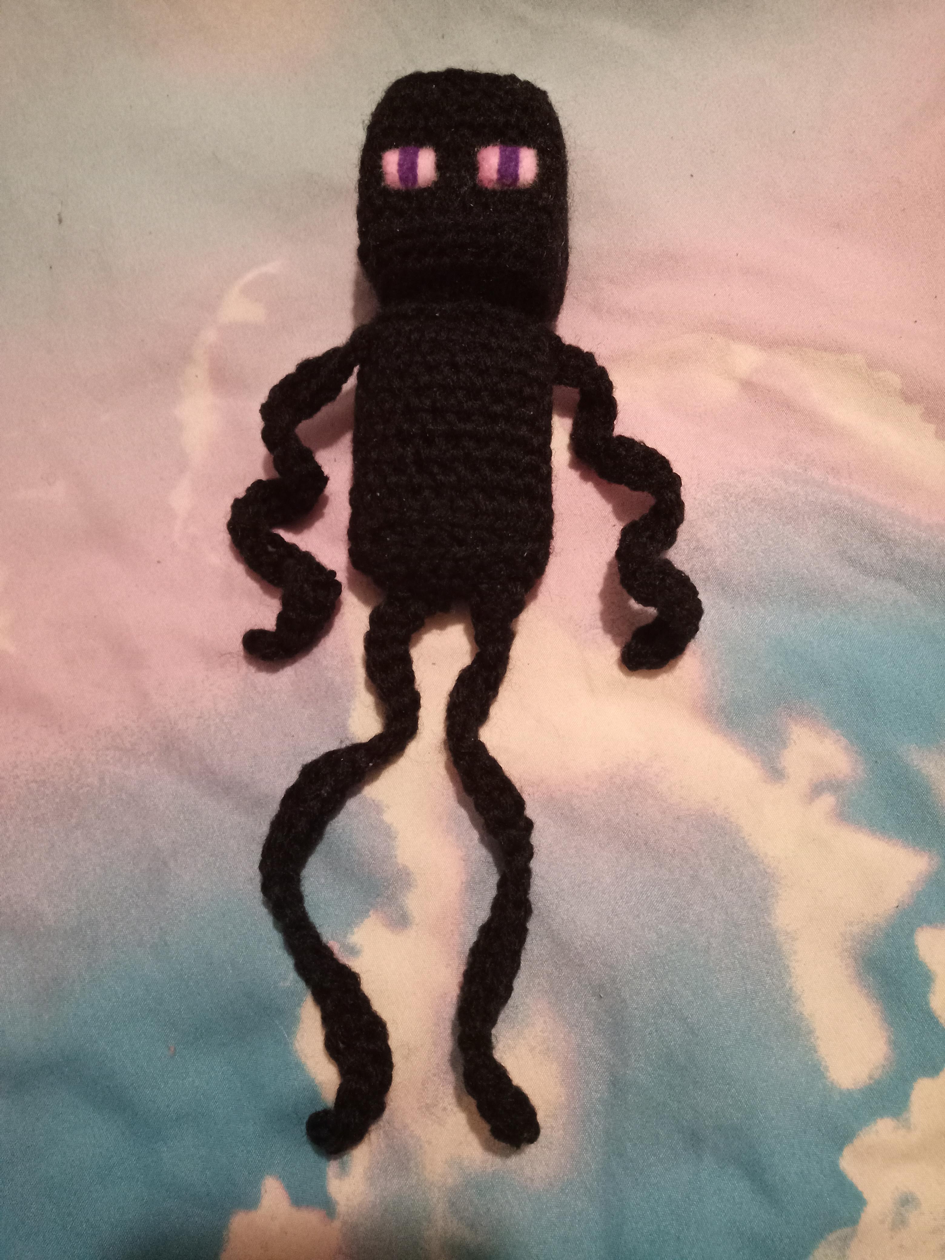 a crocheted enderman from minecraft with long curly arms and legs laying on a cloudy skies comforter