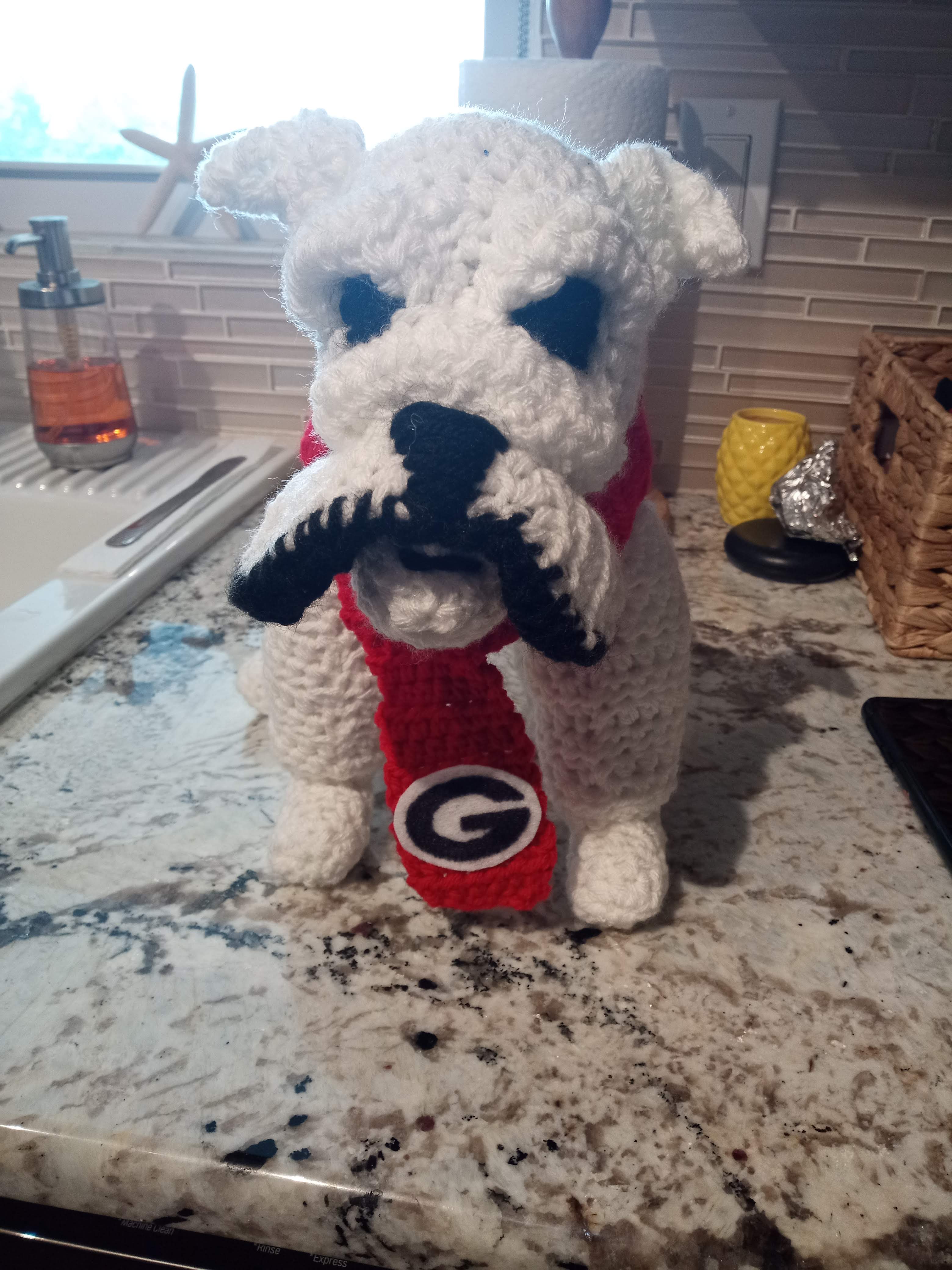a crocheted white bulldog with black eyes and a red scarf with a Georgia University logo on the end standing on a marble kitchen counter next to a sink