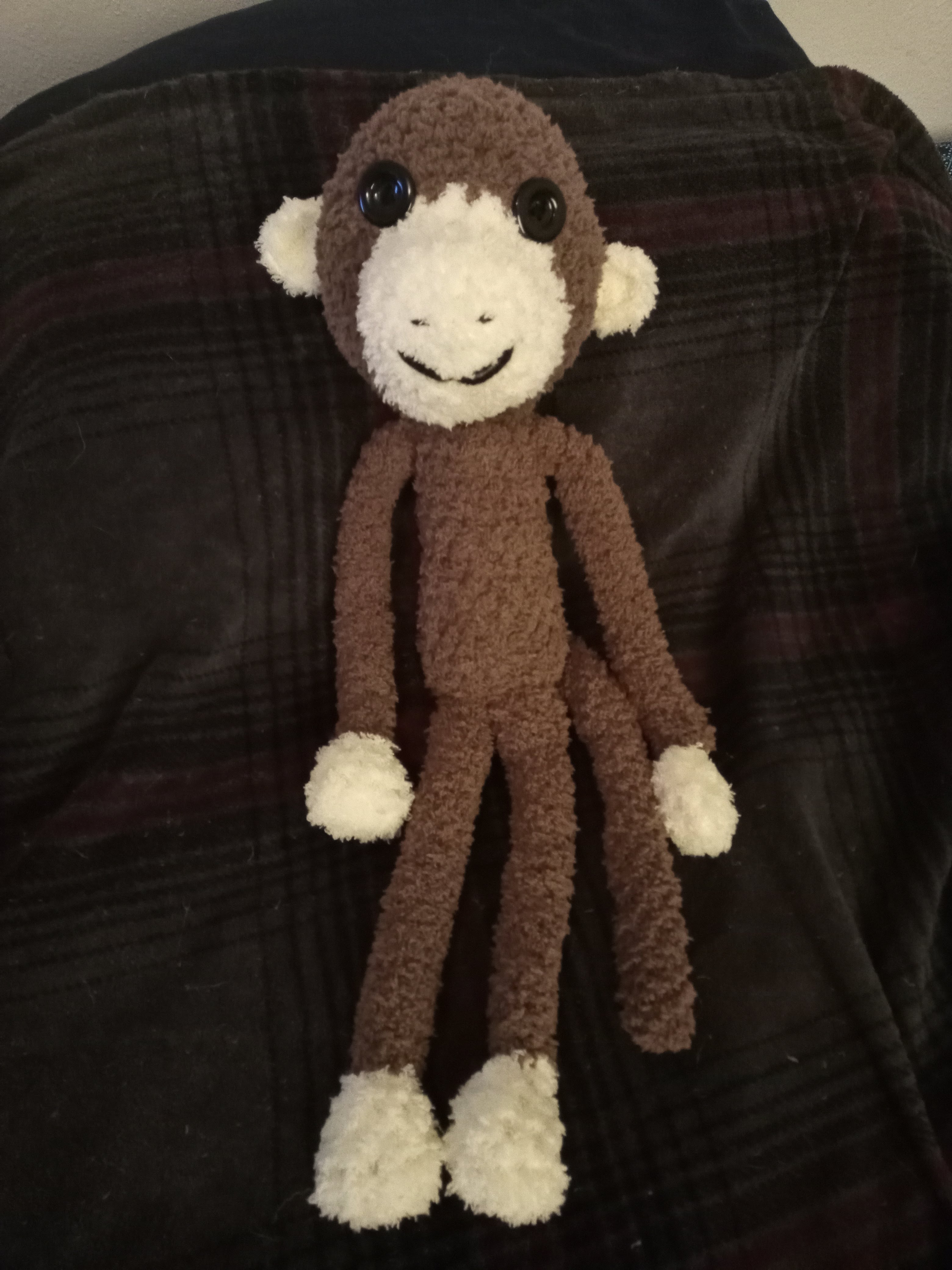 a crocheted brown and tan monkey with black button eyes laying on a brown plaid blanket