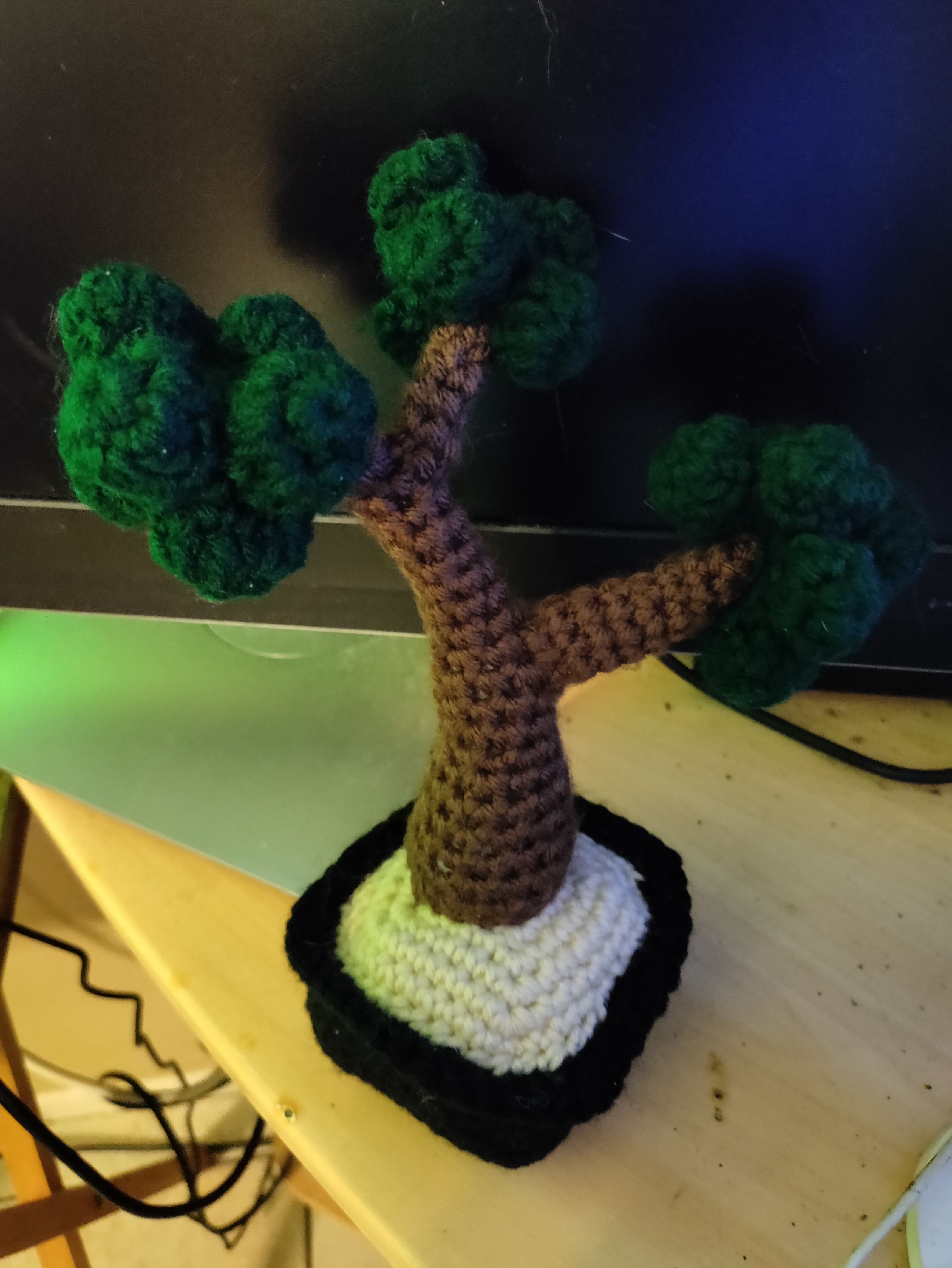 a crocheted bonsai tree in a crocheted black pot leaning against a monitor screen