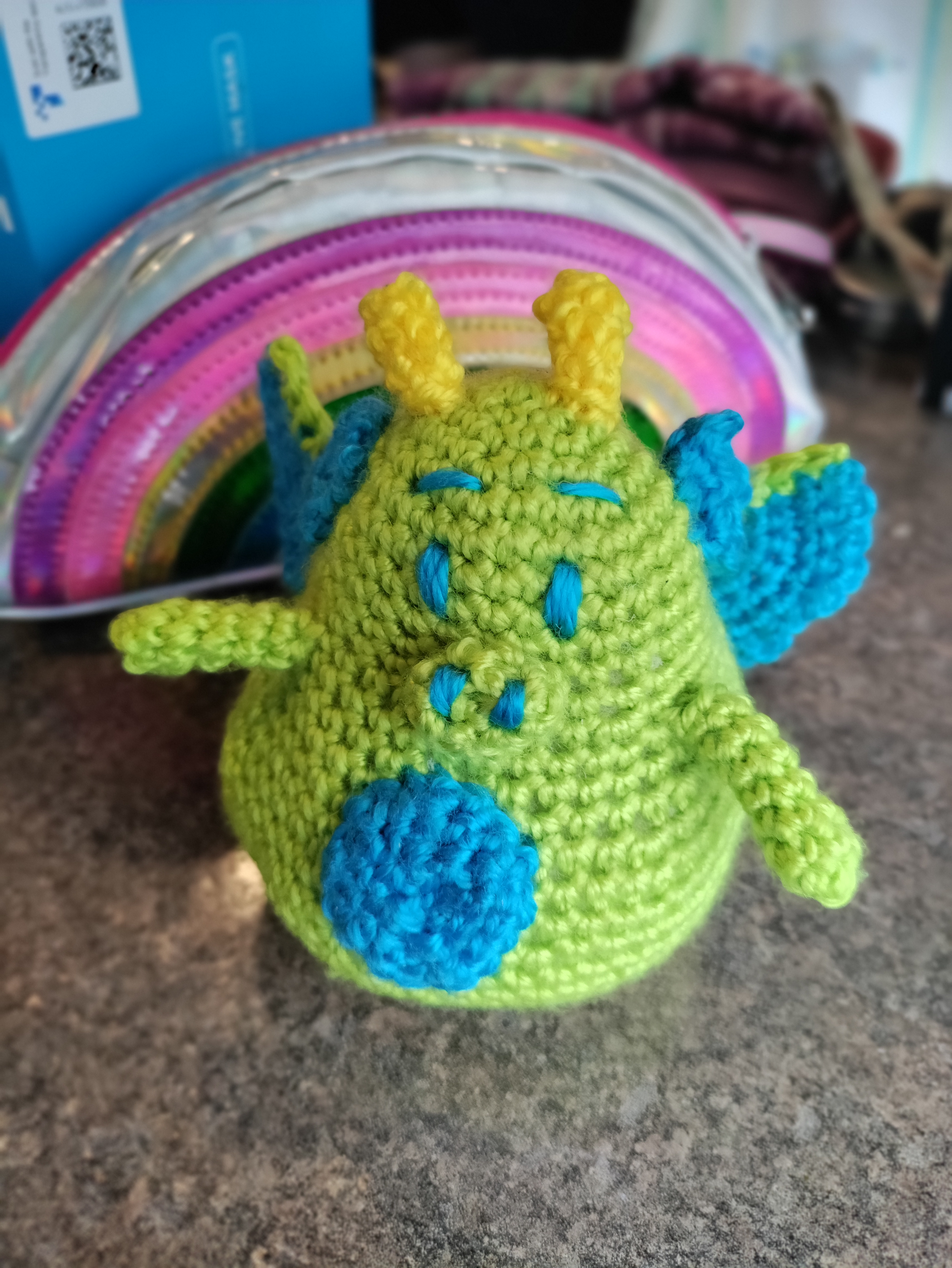 a small crocheted lime green dragon with blue eyes, eyebrows, wings, tummy patch, and nostrils on a little lime green snout as well as yellow horns standing on gray marble with a rainbow shaped pencil bag behind it