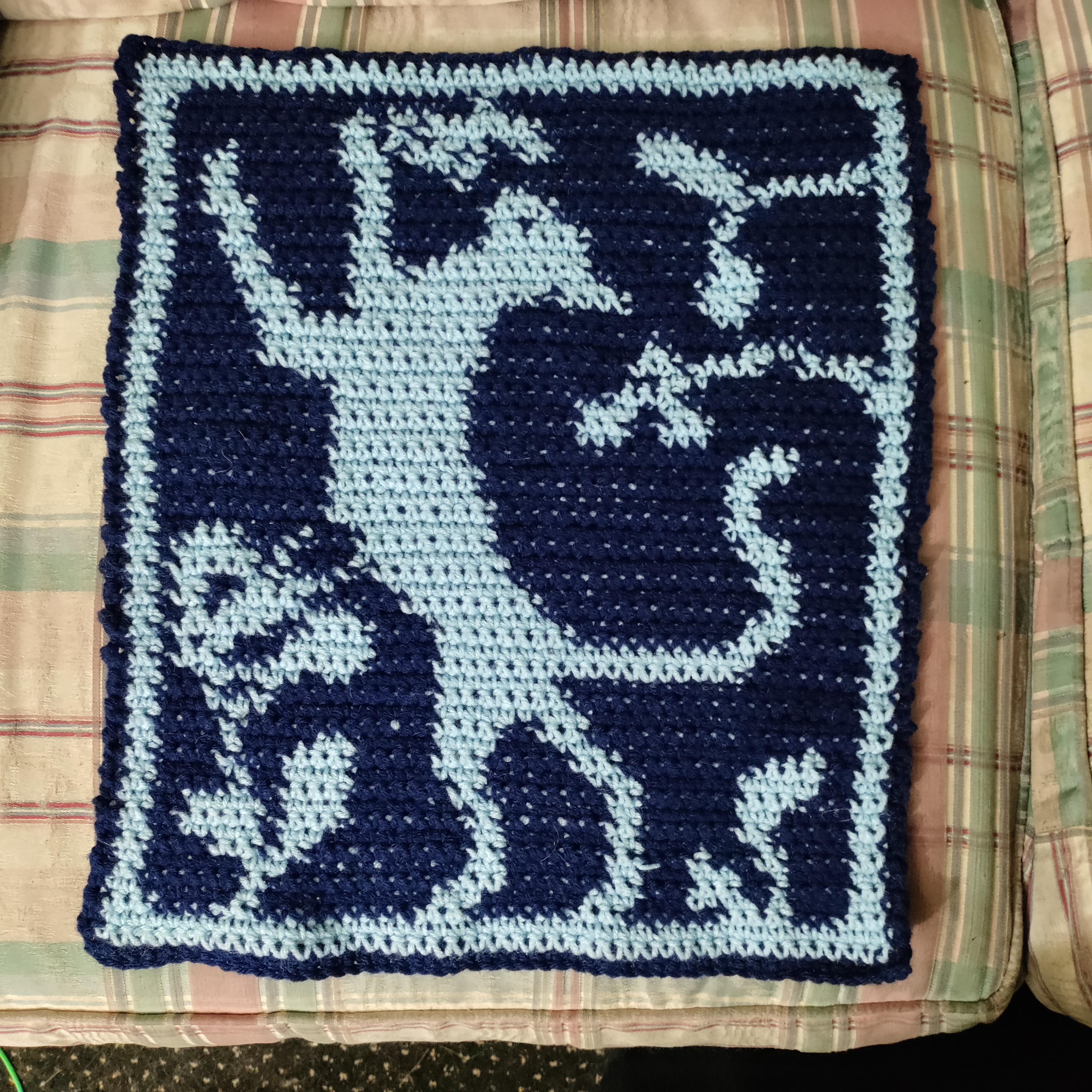 a light blue and dark blue wrinkly crocheted tapestry depicting a cat standing up on its back feet attempting to catch a butterfly next to its face with leaves and another butterfly on a flower in the background, there are gaps between stitches letting other color yarn show through