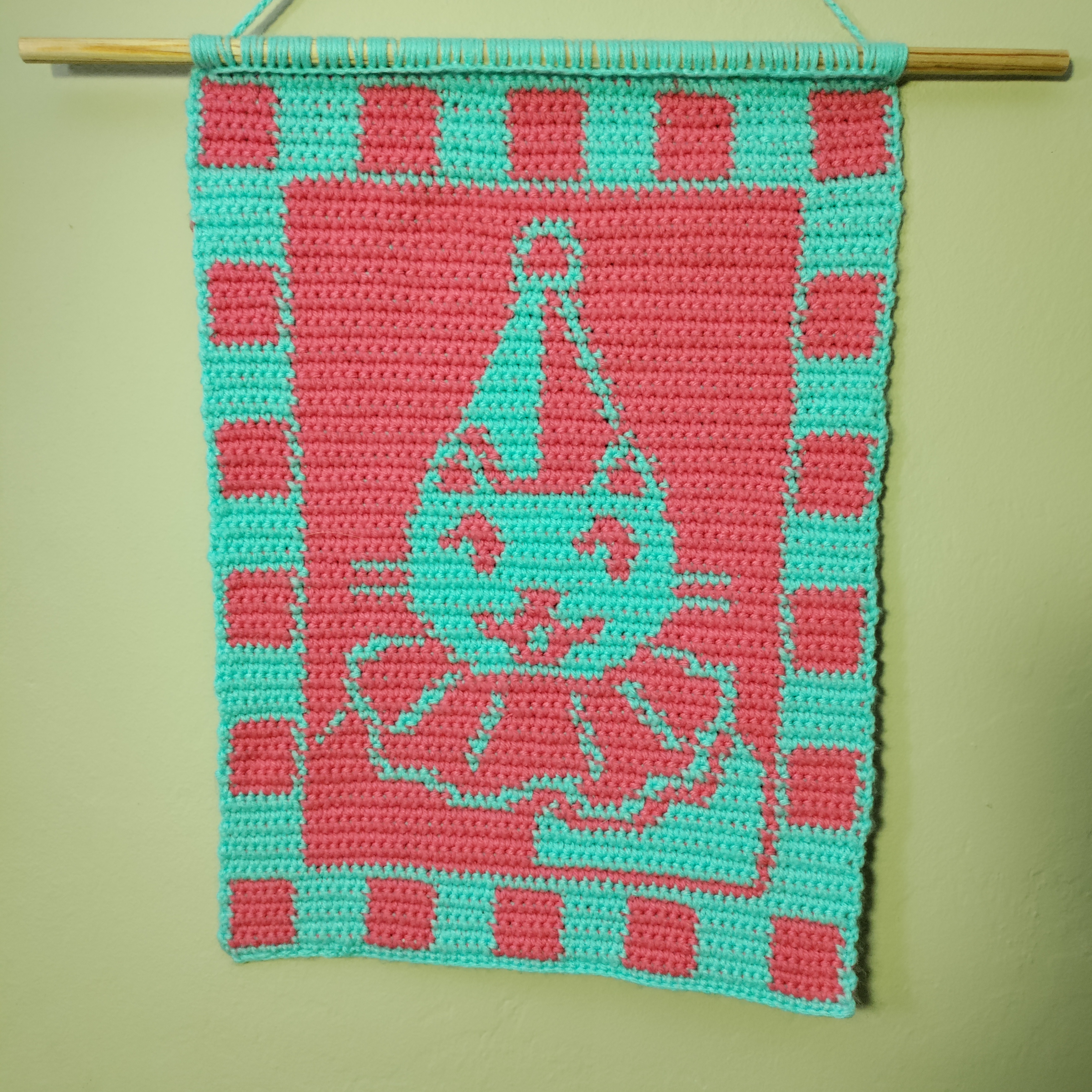 a neater looking cyan and hot pink crocheted tapestry depicting a cat in a clown outfit smiling and looking to the right, surrounded by a checkerboard border