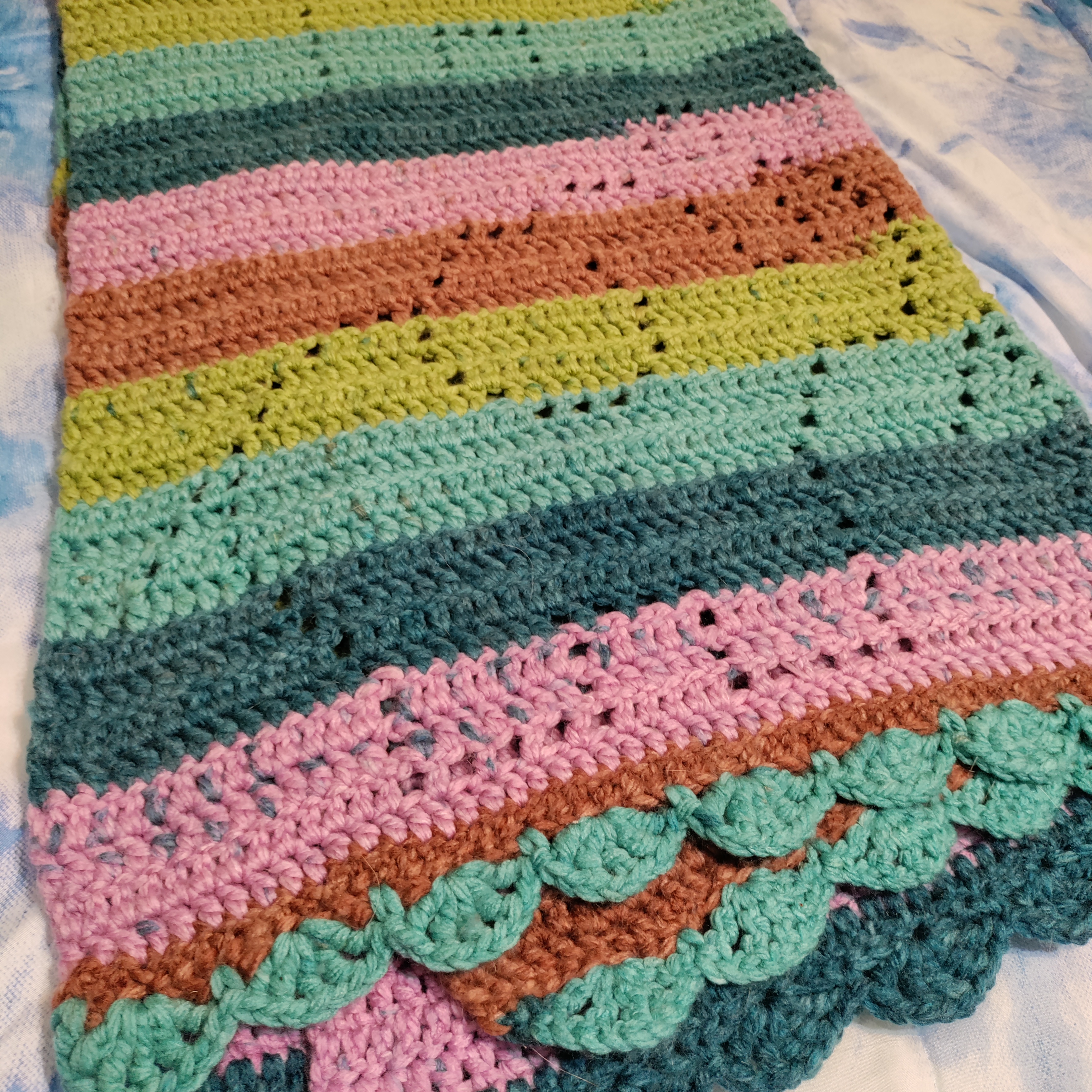 a more close up picture of a crocheted blanket with light and dark teals, olive green, brown, and pink stripes that has open stitches shaped like cute elephants with their trunks lifted and hearts above them, folded and zoomed into one of the elephants