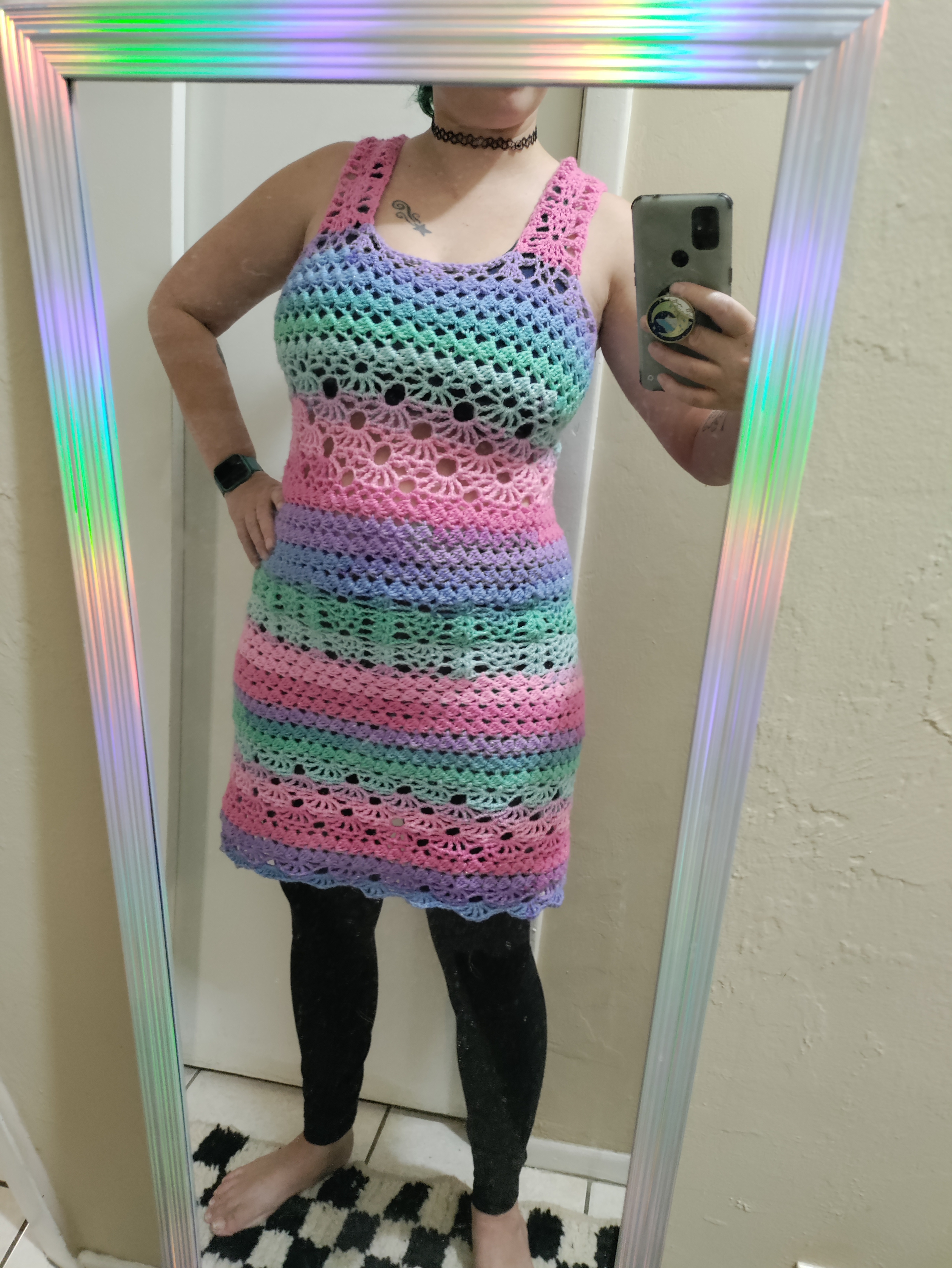 a person standing on a black and white checkered rug in front of a mirror with holographic border wearing a lacy pastel rainbow striped crocheted dress over black crop and leggings