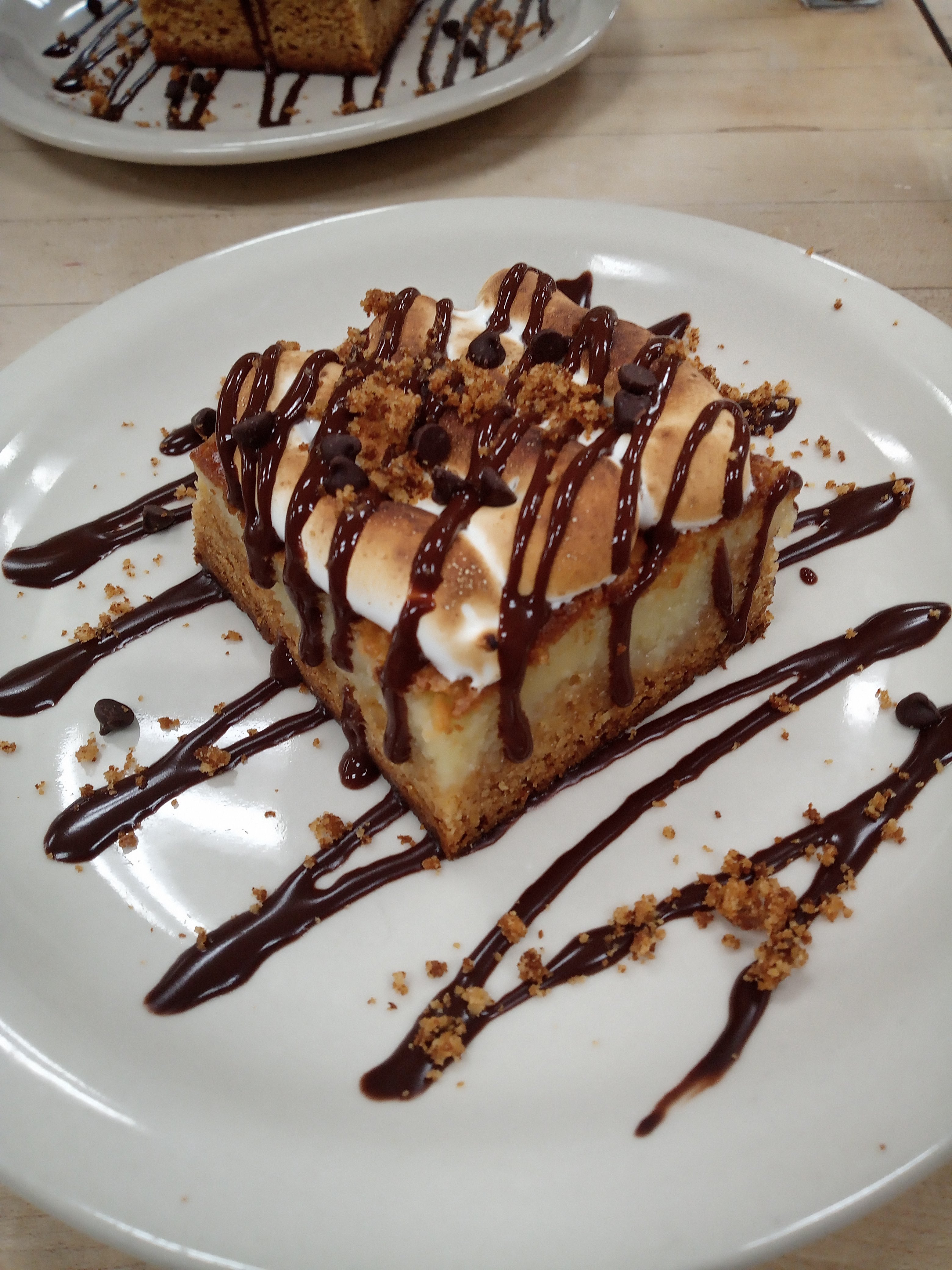 a square-cut piece of a baked dessert on a white plate topped with toasted meringue, drizzled chocolate sauce, chocolate chips, and graham cracker crumb garnish