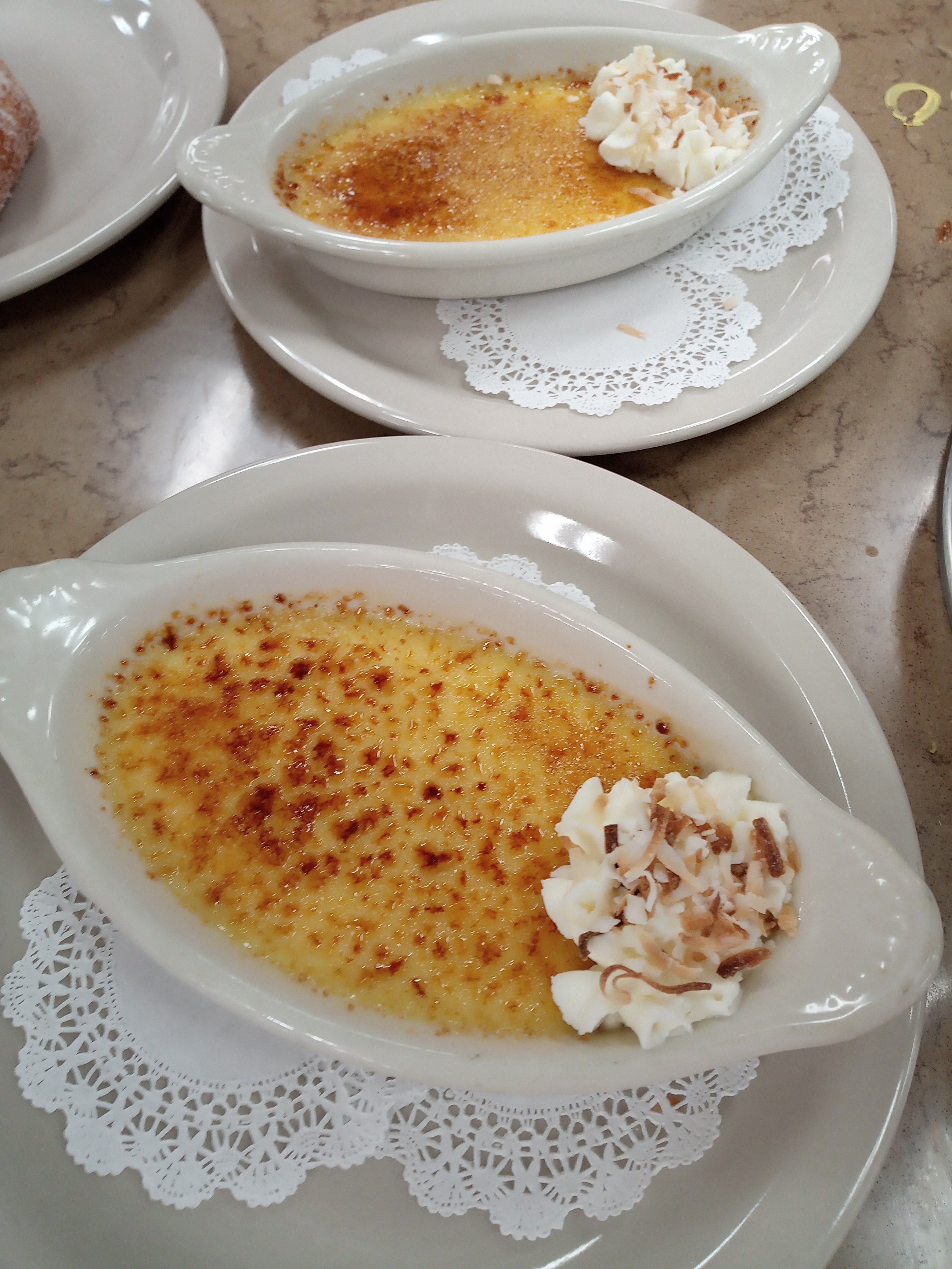 two creme brulees garnished with whipped cream and toasted coconut in oval ramekins on doily covered plates