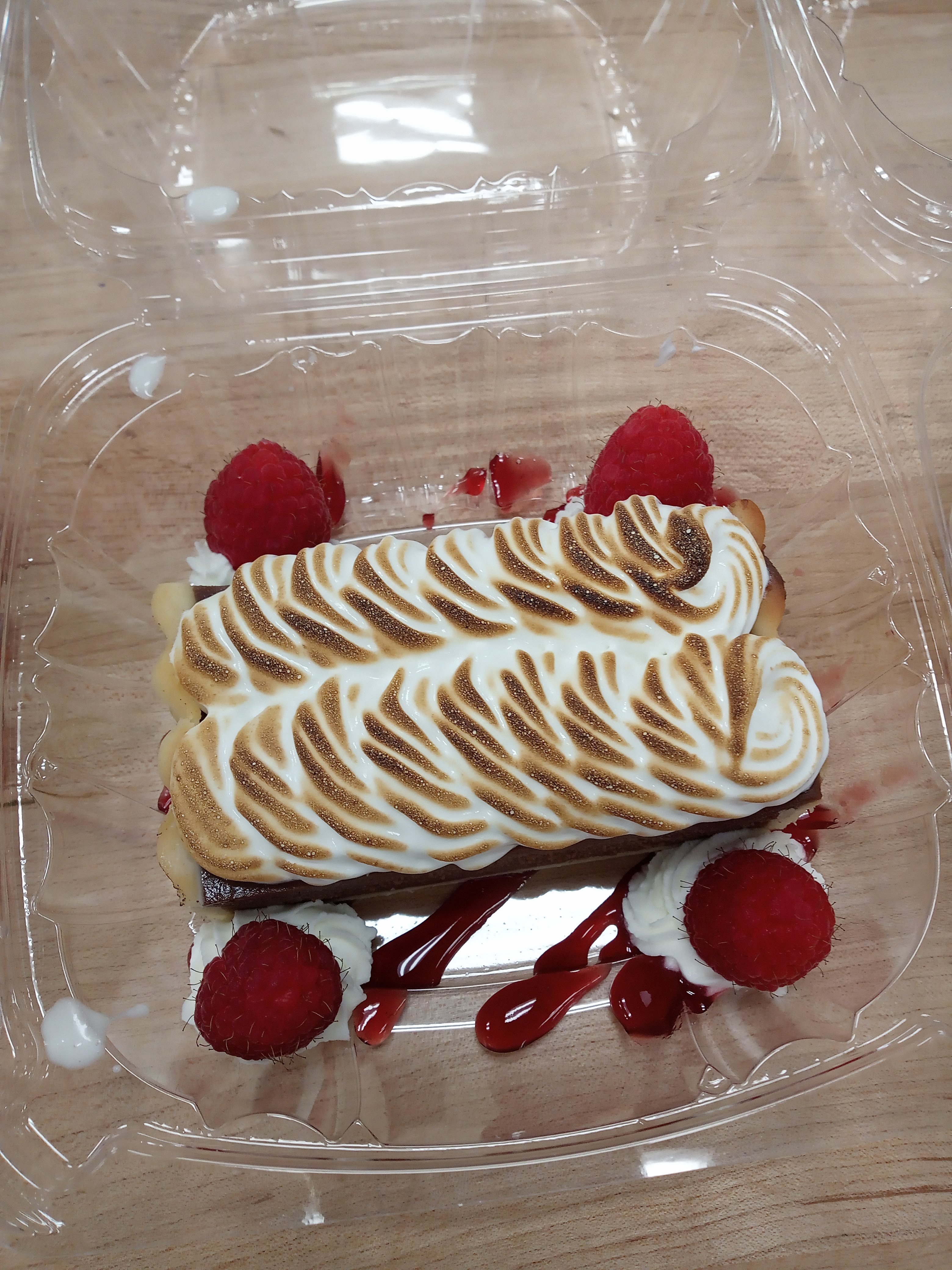 a rectangle piece of chocolate tart covered in piped toasted meringue in a clear to go container with whipped cream, drizzled red raspberry sauce, and fresh raspberries