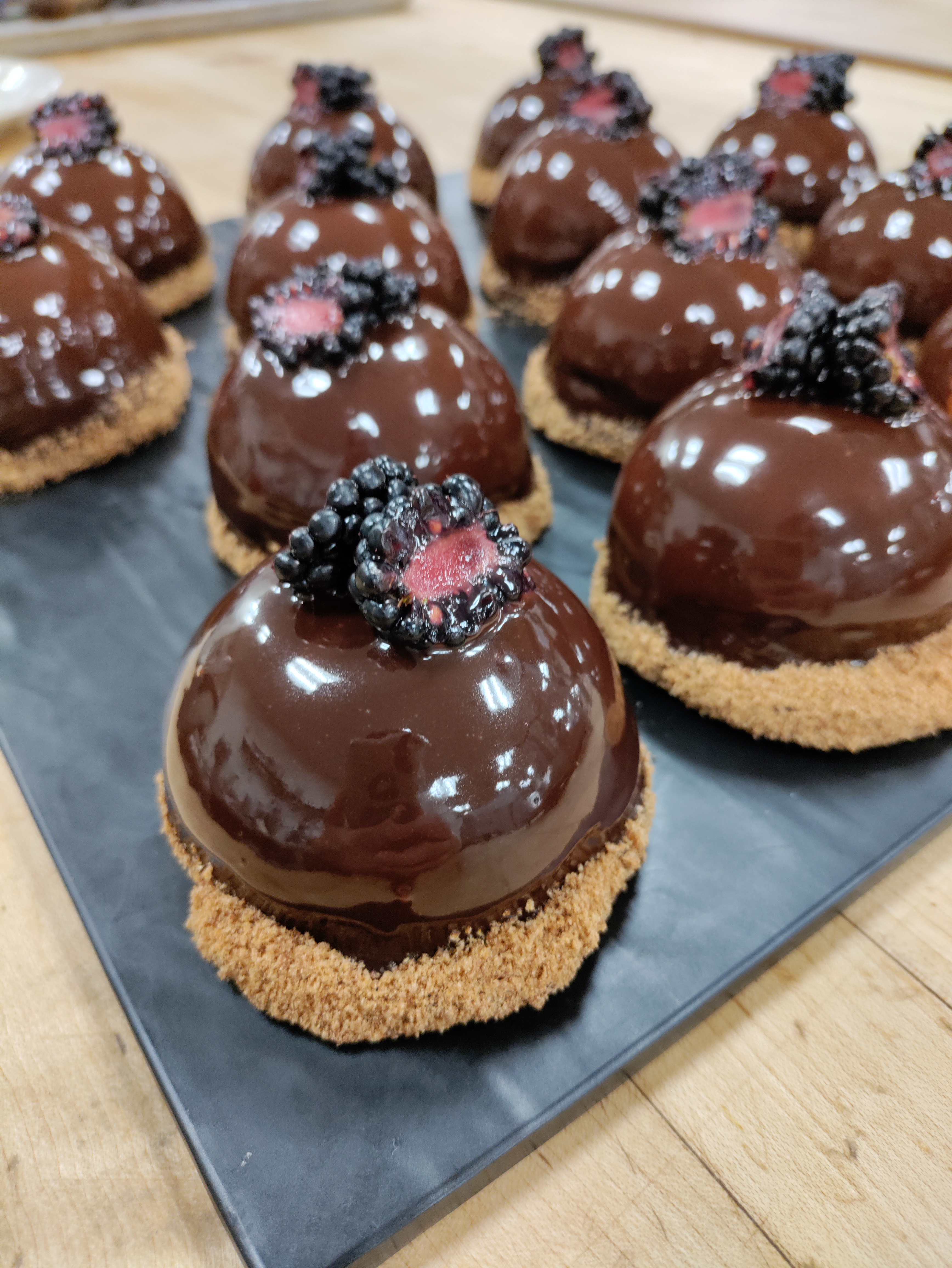 chocolate covered dome desserts topped with a split blackberry and coated with graham crumbs around the base on top of a black board