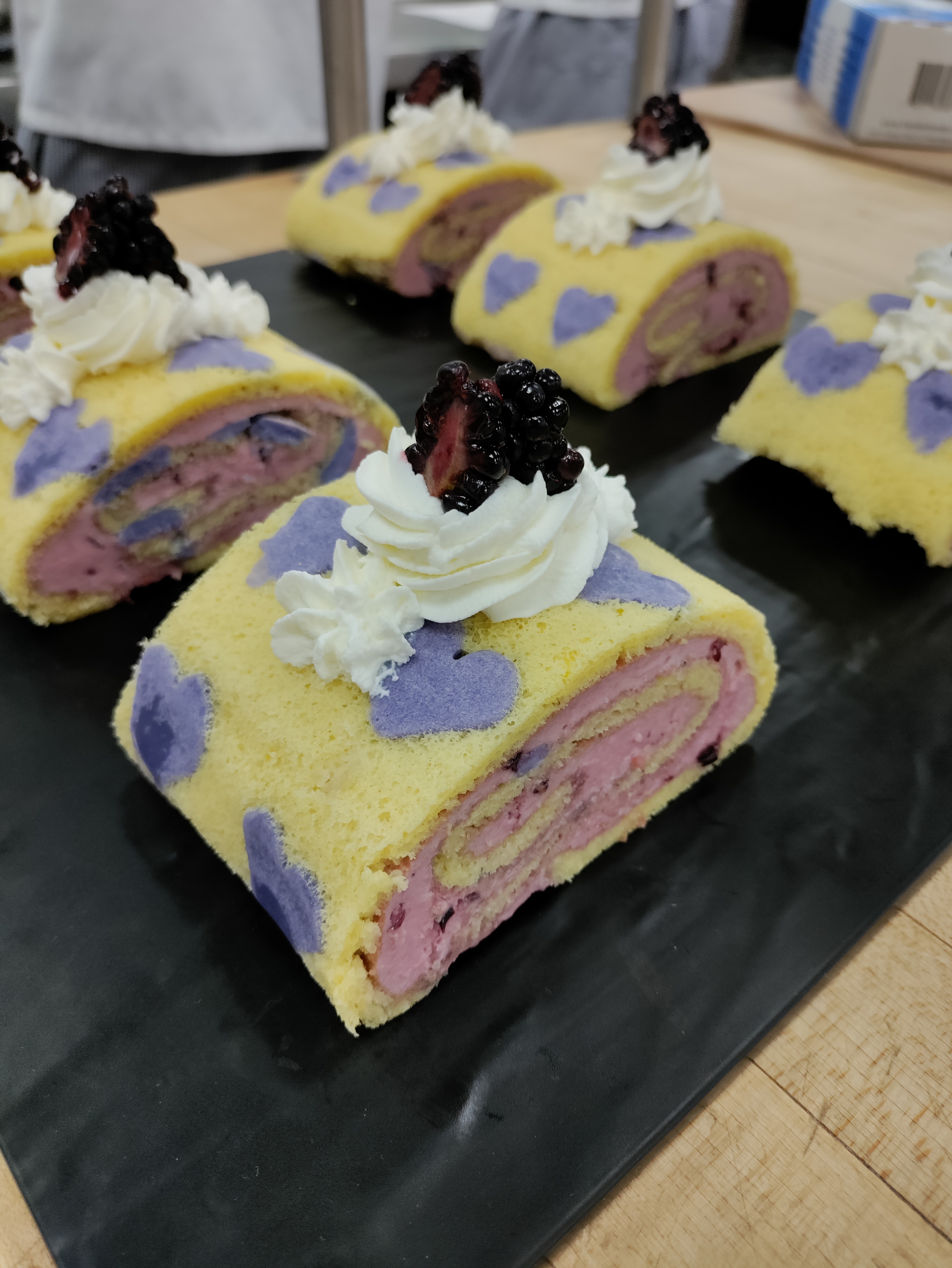 a yellow swiss roll cake lined with purple hearts and filled with a purple mousse swirl, garnished with whipped cream and two split halves of a blackberry