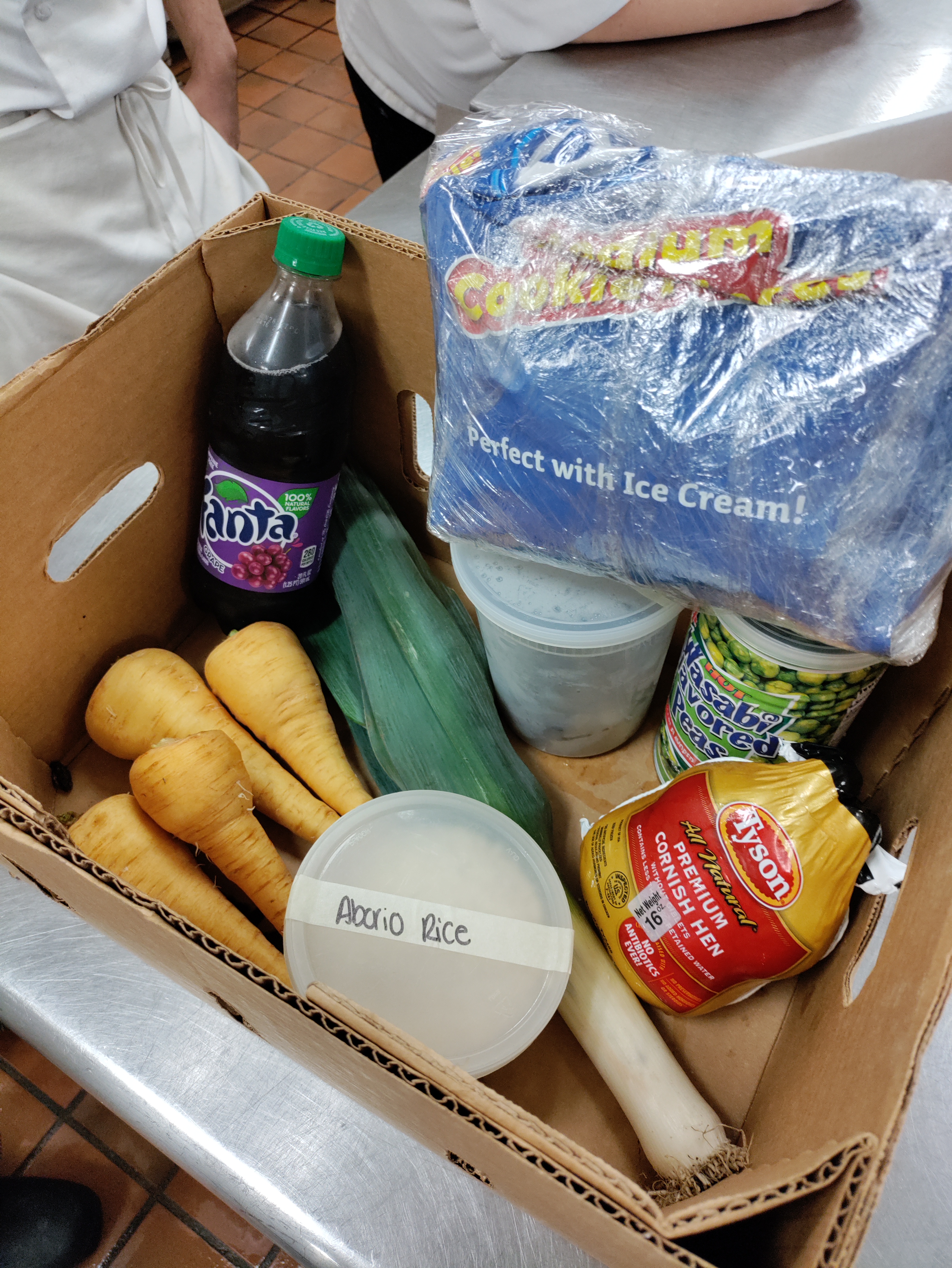 a cardboard box filled with various food items including four parsnips, one leek, a labelled container of arborio rice, a bottle of grape fanta, a cornish hen, a container of shrimp, a bag of oreo pieces, and a can of wasabi peas