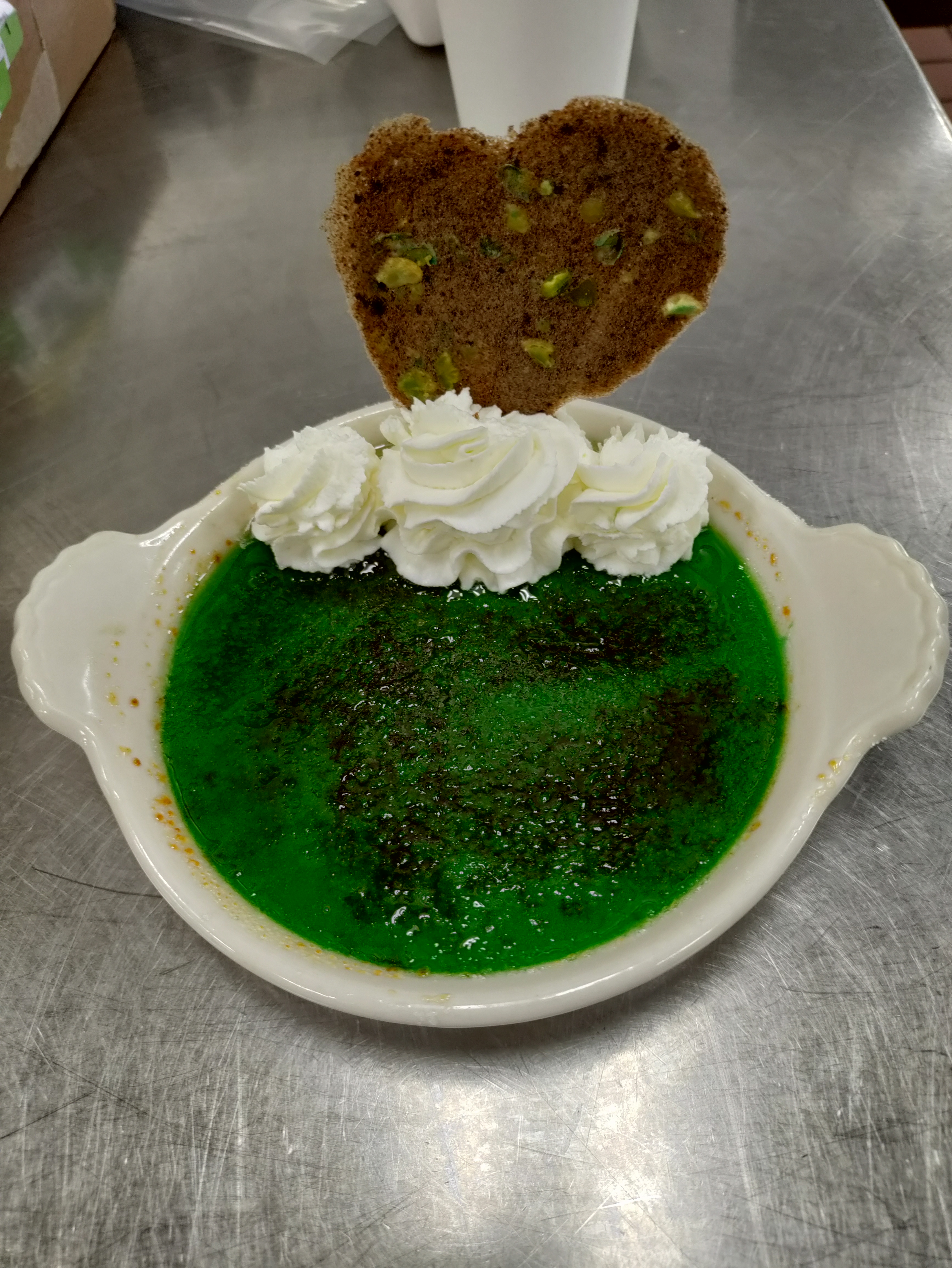 a vivid green creme brulee dessert in a ceramic ramekin garnished with whipped cream and a heart shaped tuile with visible chunks of wasabi peas and oreo dust