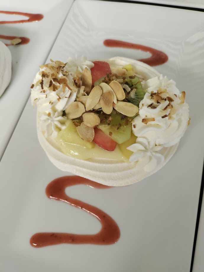 a dessert on a white rectangle plate decorated with a wiggly line of red berry sauce consisting of a white baked meringue base topped with chopped fresh fruit, whipped cream, toasted coconut and almonds